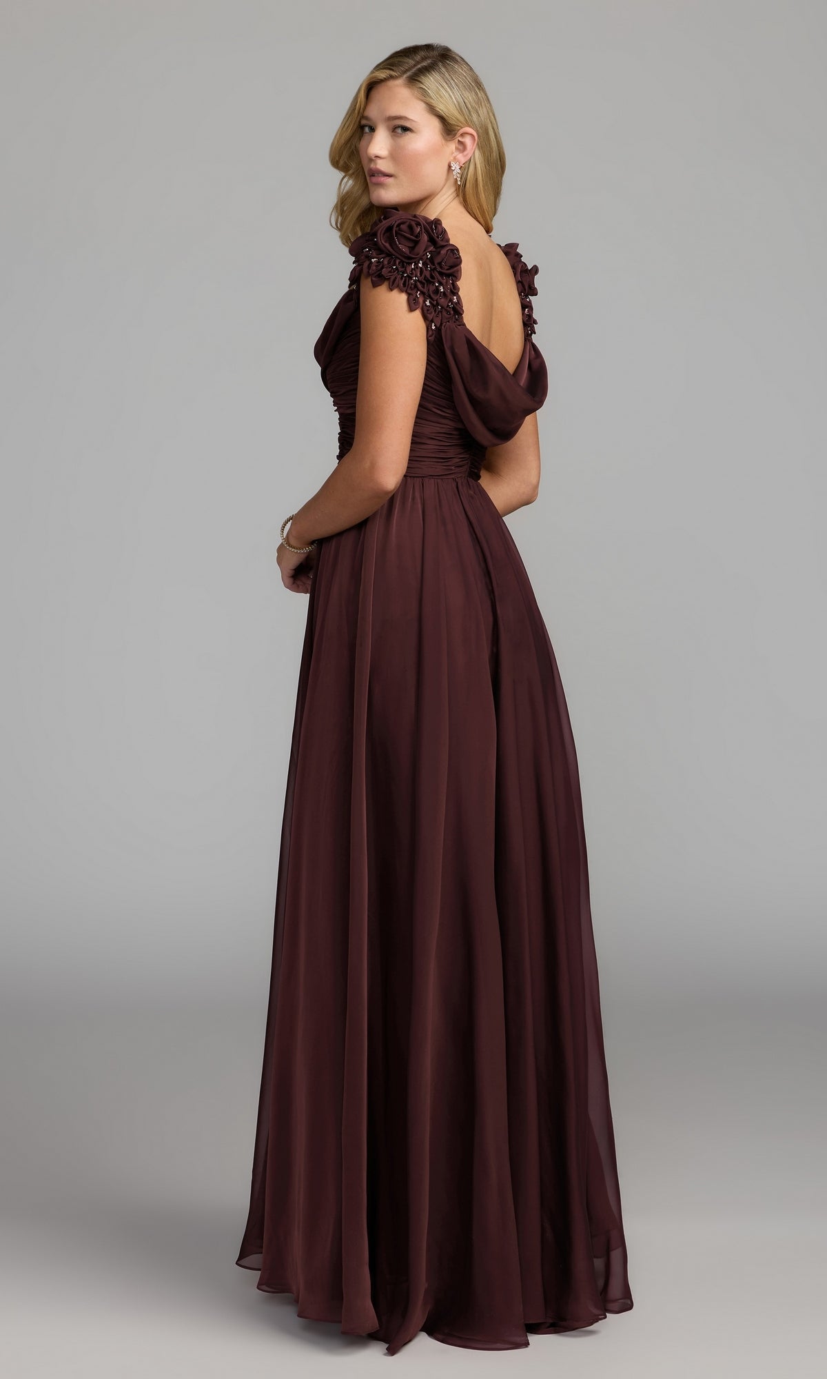 A woman stands against a plain gray background wearing the Mac Duggal 69088—a floor-length, burgundy formal dress featuring a graceful cowl neckline and floral embellishments on the shoulder.
